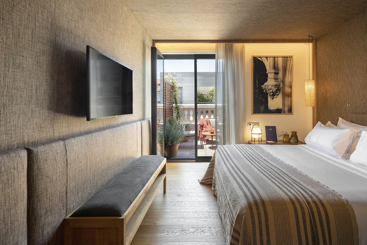 A hotel room with a large bed, a bench, a flat-screen TV mounted on the wall, and a balcony with outdoor furniture.