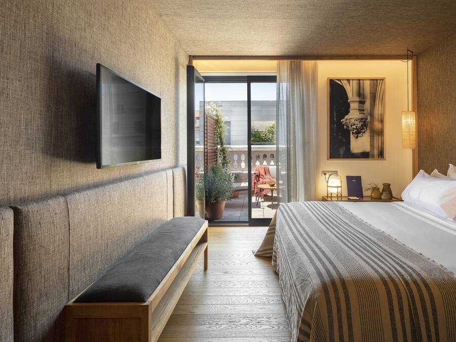 Habitación de hotel con una cama grande, una banca, televisor de pantalla plana montado en la pared y un balcón con mobiliario exterior.