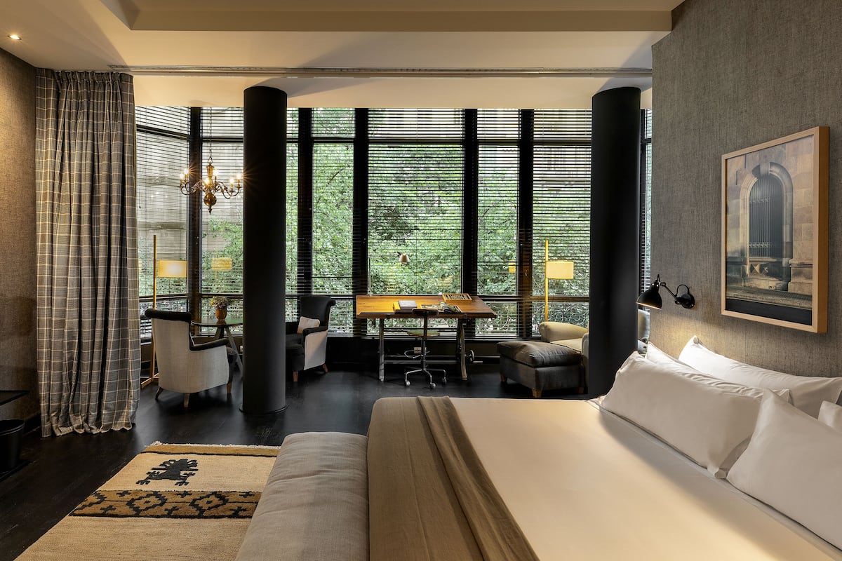 A modern hotel room with a large bed, a desk with a chair, a television, and a view through large windows.