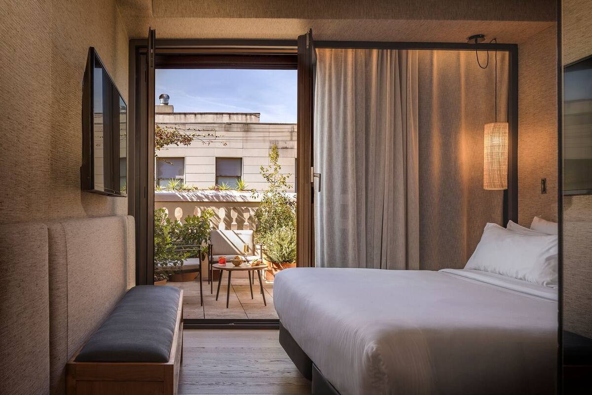 A hotel room with a bed, a bench, a table with chairs, and a view of the outside balcony.