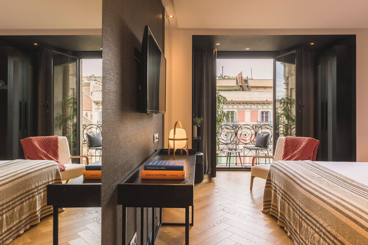 A hotel room with two beds, a desk with books, a TV, and a balcony with a view of buildings.