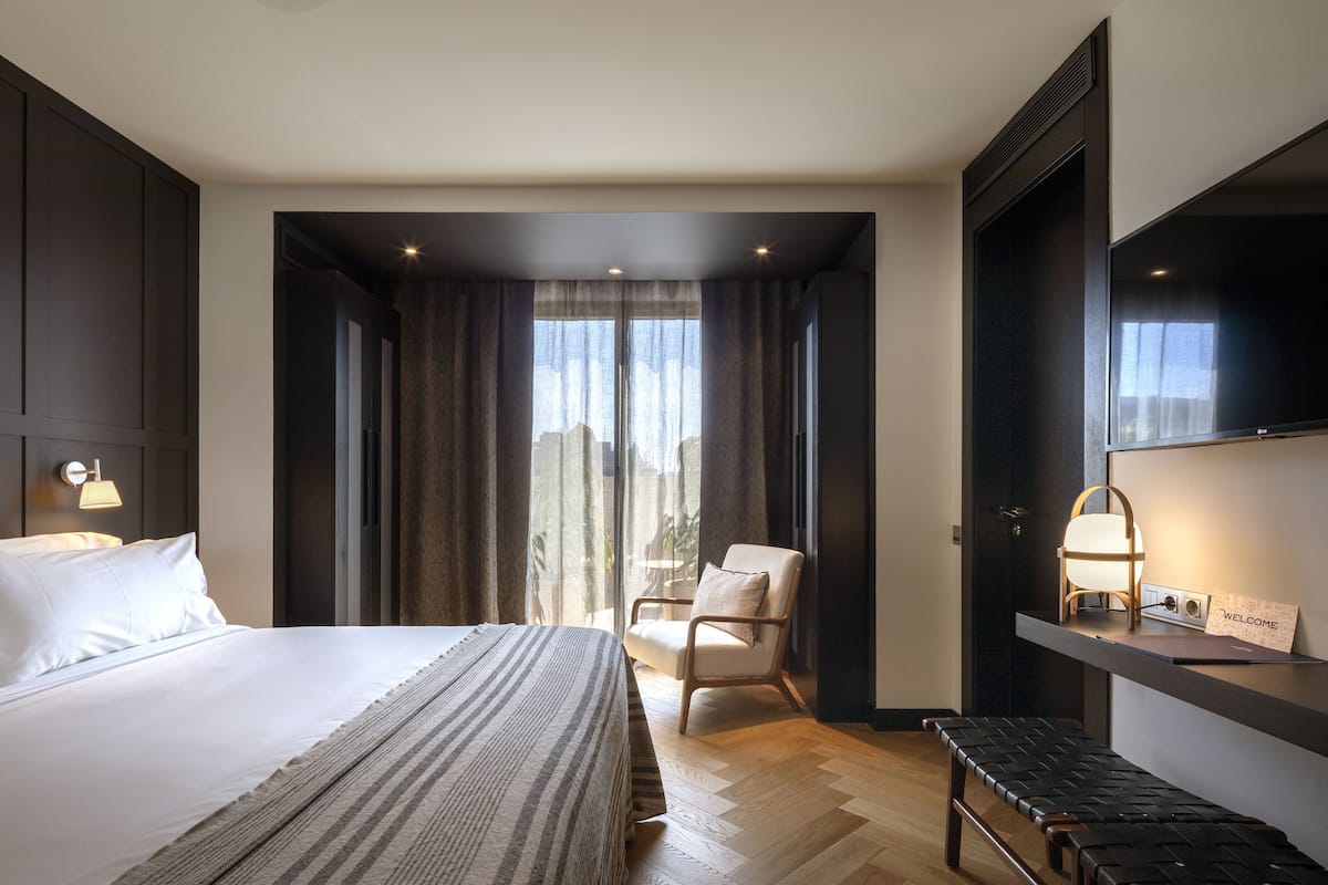 A modern hotel room with a large bed, a sitting area with a chair, a bench, and a desk with a lamp and a welcome sign.
