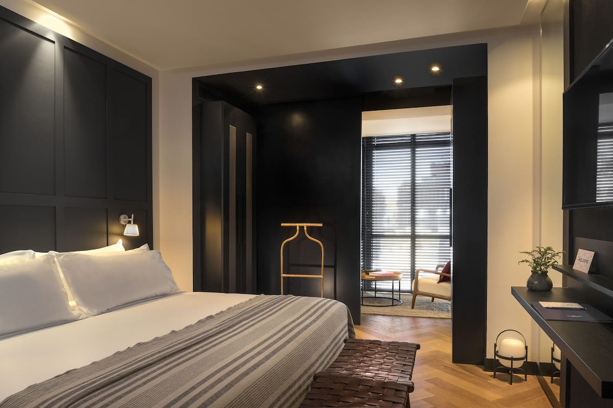 A modern hotel room with a large bed, a desk with a lamp, and a view of the outside through a window with blinds.