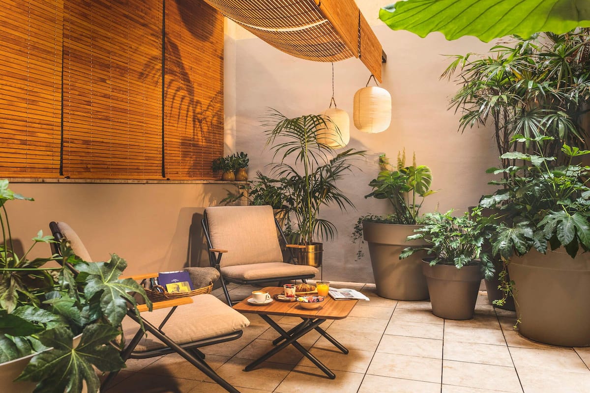 A cozy outdoor seating area with a cushioned chair, a small table with a cup and a basket, surrounded by large potted plants and a wooden ceiling structure.
