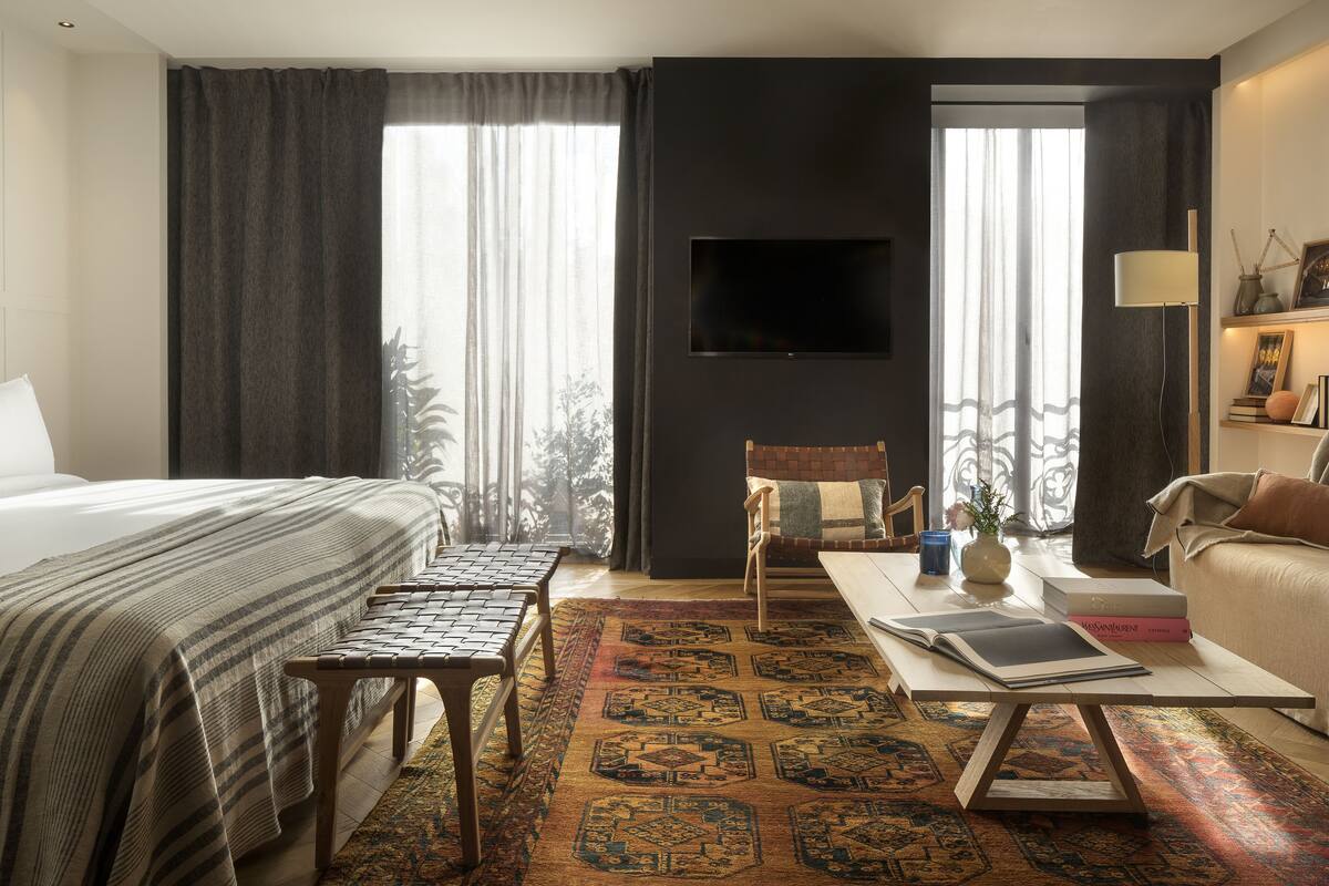 A hotel room with a bed, a sofa, a coffee table, a TV, and a view of trees through the window.
