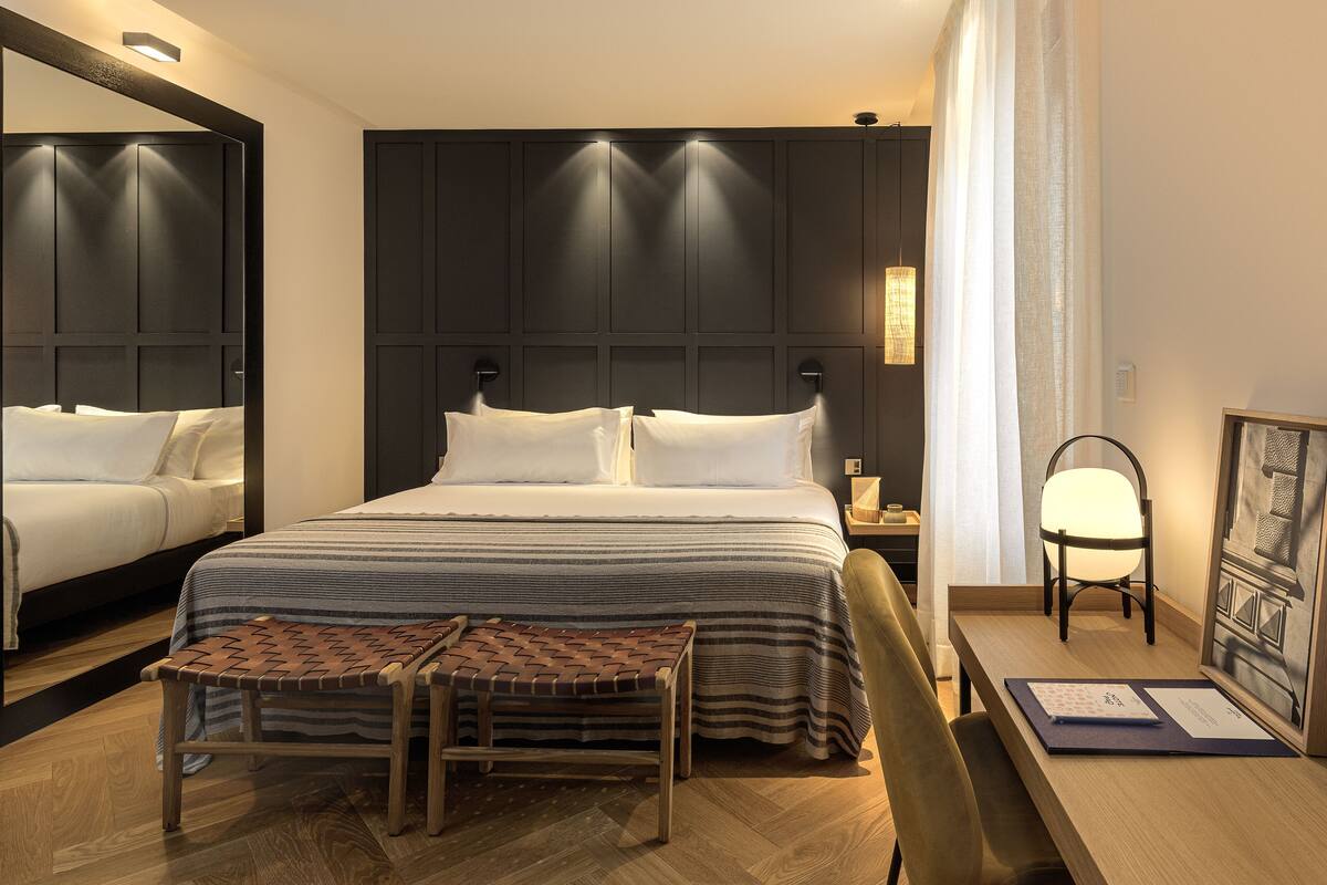 A hotel room with a large bed, a bedside table, a chair, a bench, and a framed picture on the wall.