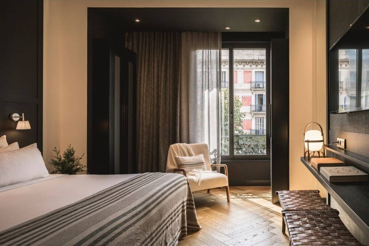 A modern hotel room with a large bed, a chair, a bench, a desk with a lamp, and a view of buildings through a window.