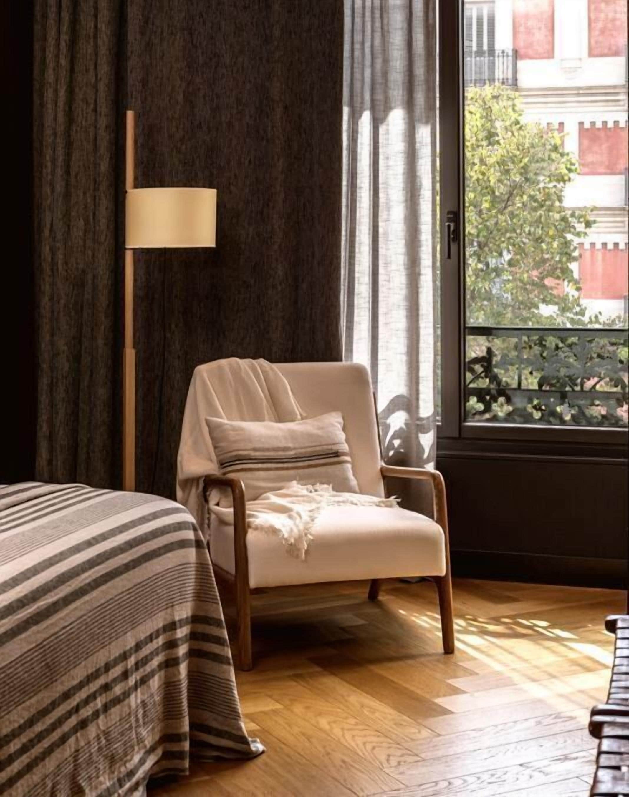 Una habitación con una cama, una silla, una lámpara de pie y una ventana con vistas a edificios y árboles.
