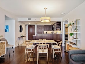 Uma área de refeições moderna com mesa e cadeiras de madeira, cozinha ao fundo e uma estante com objetos decorativos.