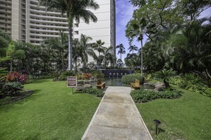 Um jardim bem cuidado com um caminho de concreto que leva a um elemento aquático, cercado por vegetação exuberante e palmeiras.