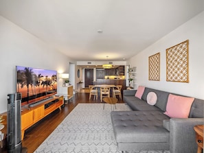 Uma sala de estar moderna com televisão de tela plana, sofá cinza, aparador de madeira e área de jantar com mesa e cadeiras de madeira.