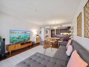 A modern living room with a flat-screen TV, a grey sofa, a wooden coffee table, and a dining area with a wooden table and chairs.