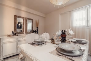 A dining area with a white table set for four, including plates, glasses, and cutlery. There are two wall mirrors with decorative frames, a vase with dried plants, and a chandelier above the table.