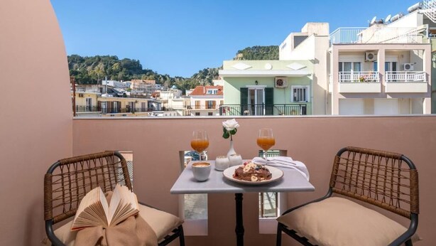 Un balcón con sillas de mimbre, una mesa con comida y bebidas, y vistas a edificios y colinas.