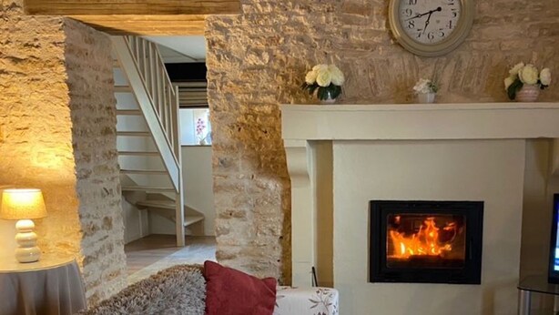 Una sala de estar acogedora con chimenea, paredes de piedra, un reloj y una escalera que sube a otro nivel.