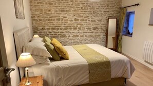 Un dormitorio con pared de piedra, una cama con cubrecama blanco y verde, una mesita de noche de madera con una lámpara, y una ventana con vistas.