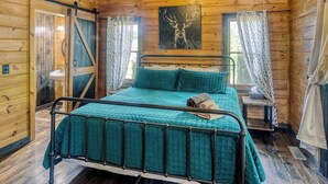 Ein Schlafzimmer einer Holzhütte mit Metallbett, blaugrüner Bettwäsche, einem Bild eines Rehs an der Wand und Vorhängen an den Fenstern.
