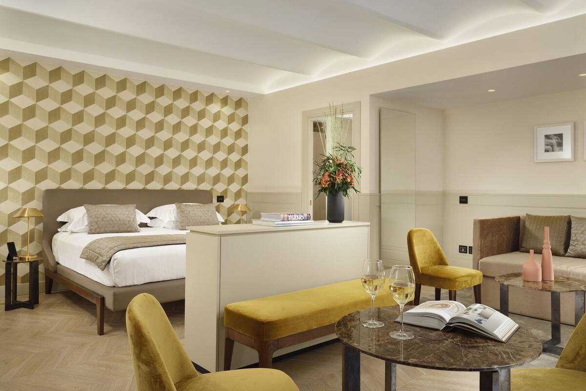 Una habitación de hotel moderna con cama, sofá, banqueta, una mesita y un jarrón decorativo.