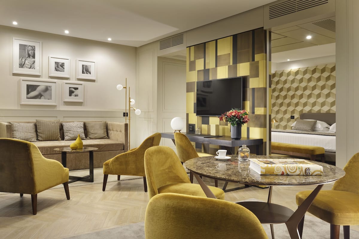 Una habitación de hotel moderna con sofá, sillones amarillos, una mesa redonda, un televisor de pantalla plana y obras de arte en las paredes.
