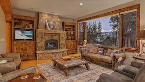 A cozy living room with a stone fireplace, wooden furniture, and a large window offering a mountain view. - Mountain Views + Hot Tub + Pet Friendly + Ski Shuttle - Alpenglow Peaks Lodge (Breckenridge)