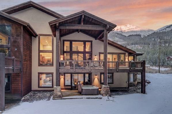 A multi-story lodge with a balcony, large windows, and a hot tub in a snowy setting.