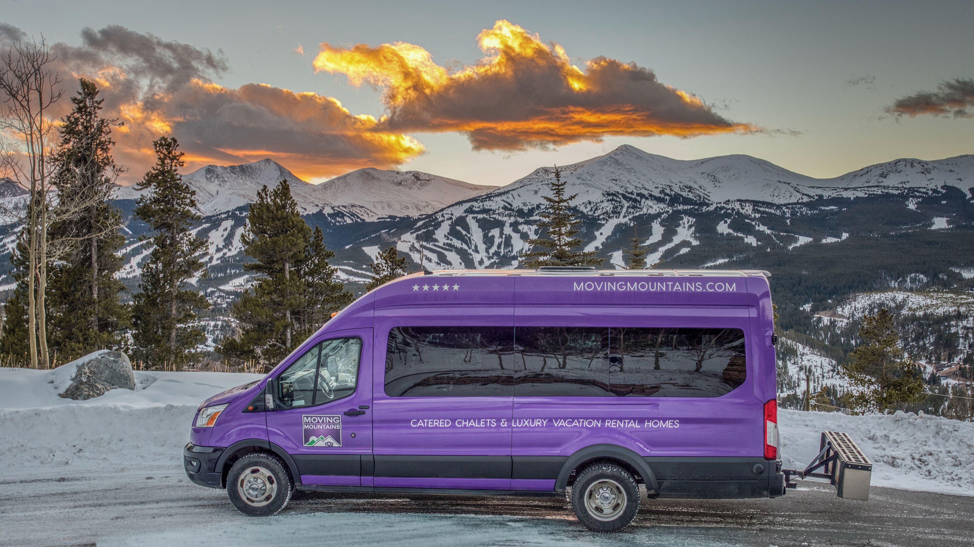 A purple moving van parked on a snowy road with mountains in the background.
