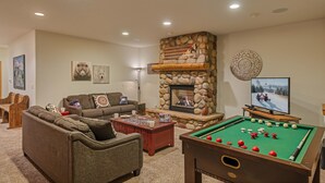 Ein gemütliches Wohnzimmer mit Kamin aus Stein, Billardtisch, einer Couch und einem Fernseher, der eine verschneite Szene zeigt.