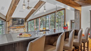 A modern kitchen with a large island, stone fireplace, and large windows overlooking a forest. - Stunning Views | Grand Vista Lodge (Breckenridge)