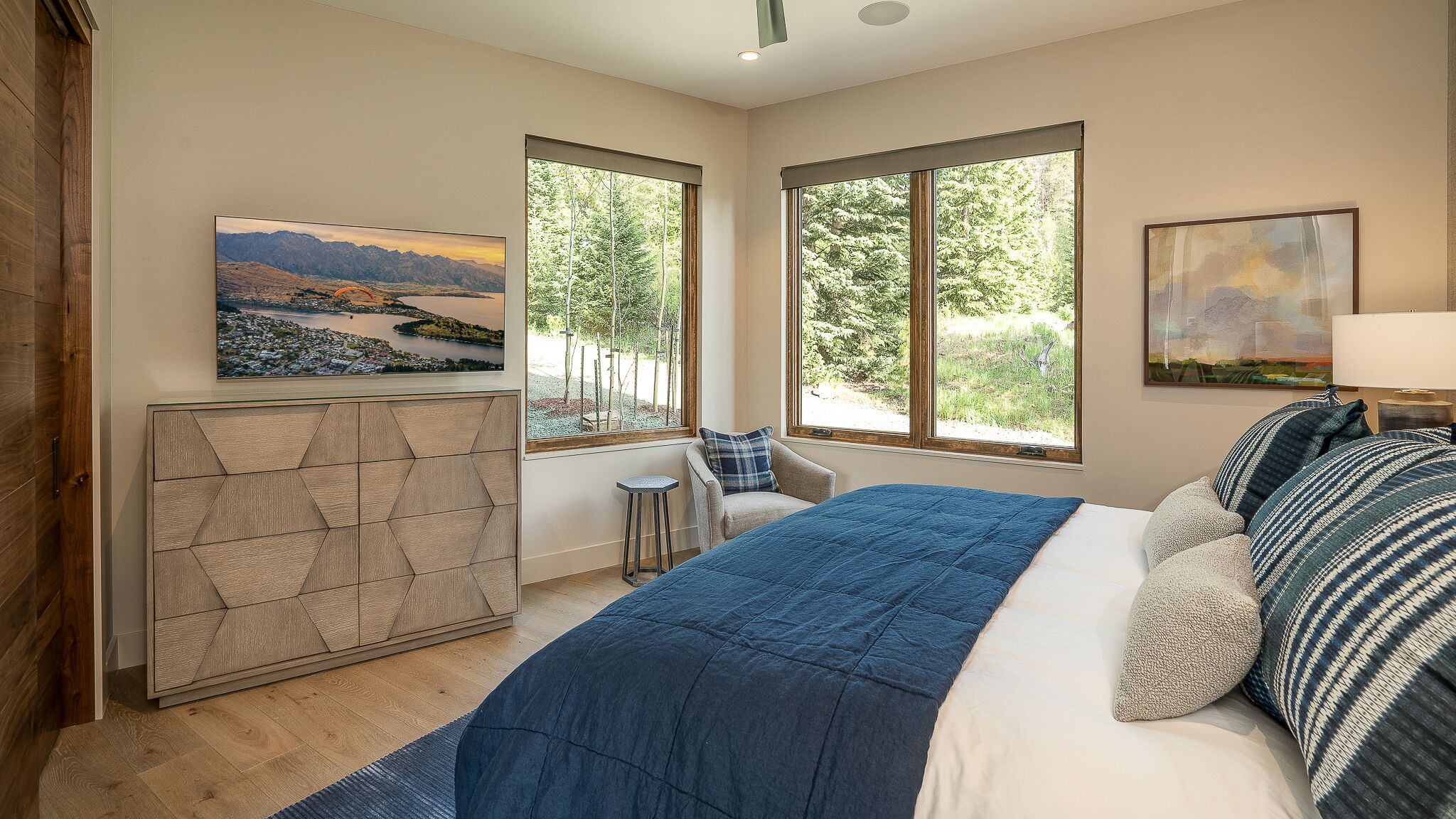 A bedroom with a large bed, two windows with a view of trees, a painting on the wall, and a cabinet with a geometric design.