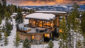 Un moderno lodge de varios niveles con terraza acristalada y bañera de hidromasaje, rodeado de árboles nevados y montañas.