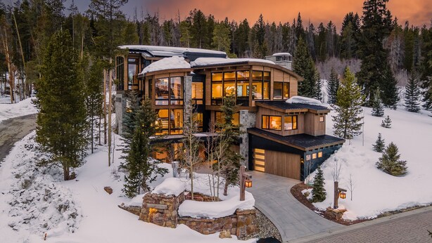 Una cabaña de madera moderna de varios pisos con grandes ventanales y cimientos de piedra, rodeada de nieve y árboles.