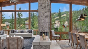 A spacious living area with a fireplace, large windows, and a view of a forested hillside. - Breathtaking Ski-in/Ski-out Home + Mtn Views + Hot Tubs - The Bear’s Den (Breckenridge)