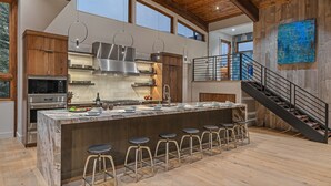 Una cocina moderna con una isla grande, taburetes de bar, electrodomésticos de acero inoxidable y una escalera que sube a un nivel superior.