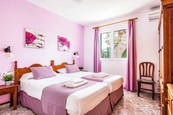 A bedroom with two beds, a wooden headboard, a window with curtains, a chair, and a small table with a plant.