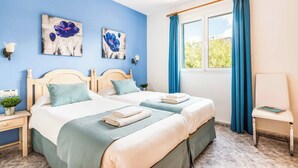 A bedroom with two beds, a window with blue curtains, and artwork on the wall.