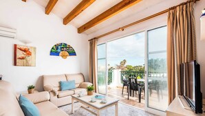 A modern living room with a beige sofa, a coffee table, and a TV on a wooden stand. There is a sliding glass door leading to an outdoor patio with a table and chairs.