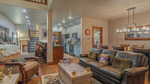 A spacious living room with a large brown leather sofa, wooden coffee table, and a jigsaw puzzle. A kitchen with blue cabinets and a dining area with a chandelier are visible in the background.
