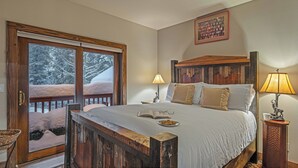A bedroom with a large bed, two bedside lamps, a wooden headboard, a framed picture on the wall, and a window with a view of snow-covered trees.