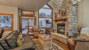 A cozy living room with a stone fireplace, leather sofa, and a large window overlooking a snowy deck.