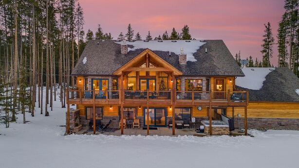 A log cabin with a balcony and a hot tub, surrounded by snow and trees.