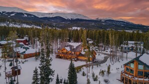 A log cabin with a large deck and multiple windows, surrounded by snow-covered ground and trees, with mountains in the background.