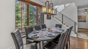 Un espace repas avec une table ronde dressée pour six personnes, entourée de fauteuils en cuir, d’un escalier et offrant une vue sur les arbres à travers de grandes fenêtres.