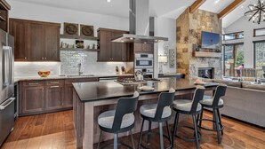 Une cuisine moderne avec des meubles de couleur bois foncé, un grand îlot central doté de tabourets, un mur d’accent en pierre et une cheminée.