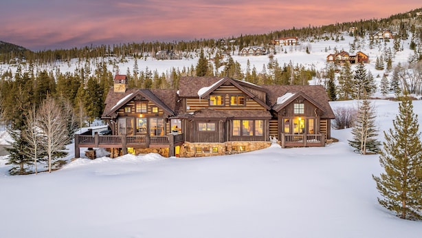 A large, multi-story log cabin with a stone foundation, multiple balconies, and large windows, surrounded by a snowy landscape and dense forest.
