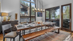 Un comedor con una larga mesa de madera dispuesta para seis personas, rodeada de sillas. Hay un banco en el centro, y la habitación cuenta con ventanas grandes y vistas al exterior.