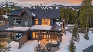 Una cabaña de troncos moderna con una amplia terraza, jacuzzi y chimenea, rodeada de montañas cubiertas de nieve.