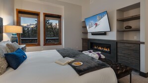 A modern bedroom with a large bed, a fireplace, a television, and a view of a snowy landscape through the windows. - Spacious w/Mountain Views + Hot Tub + Close to Skiing - Mountain Gate (Breckenridge)