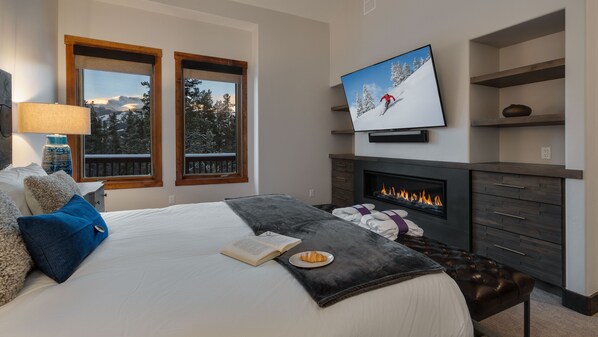 A modern bedroom with a large bed, a fireplace, a television, and a view of a snowy landscape through the windows. - Spacious w/Mountain Views + Hot Tub + Close to Skiing - Mountain Gate (Breckenridge)