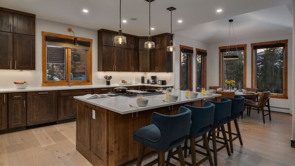 A modern kitchen with dark wood cabinets, a large island with bar stools, and large windows offering a view of the outdoors. - Spacious w/Mountain Views + Hot Tub + Close to Skiing - Mountain Gate (Breckenridge)