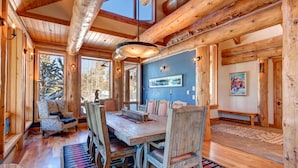 A rustic wooden interior with a large dining table, wooden chairs, and a bench. There are large windows allowing natural light and views of the outdoors.